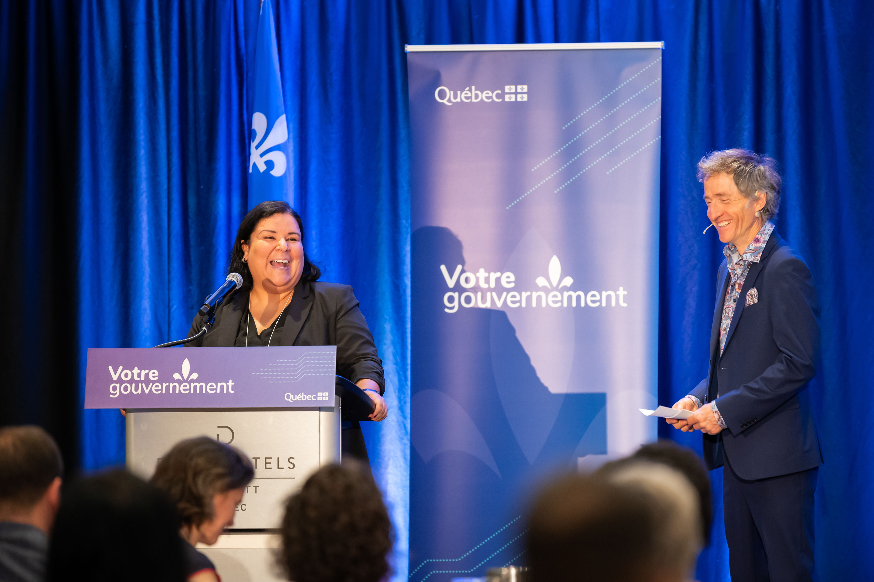 Tanya Sirois, du Regroupement des centres d'amitié autochtones du Québec, présente au lutrin en riant avec Emmanuel Bilodeau.